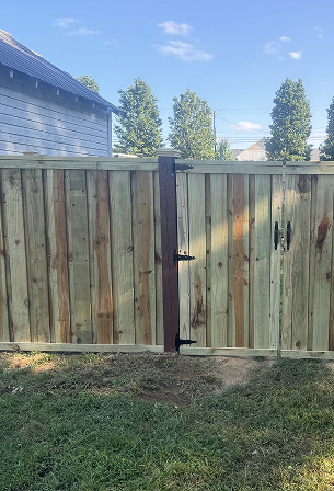 Wooden backyard fence with a gate section, set on a grassy area with house and trees in the background under a blue sky.