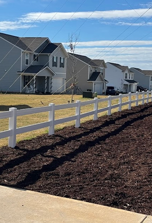 Row of modern suburban houses with white vinyl fences and a fresh mulch bed under a blue sky.