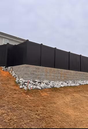 Black vinyl privacy fence installed on top of a stone retaining wall with white rocks and brown grass on a cloudy day.