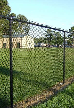 Black chain-link fence surrounding a grassy area with a beige building and trees in the background under a clear blue sky.