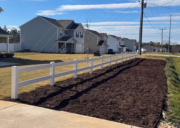 Freshly mulched garden bed bordered by a white vinyl fence along a suburban street with houses and utility poles.
