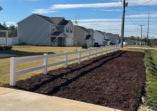 Freshly mulched garden bed bordered by a white vinyl fence along a suburban street with houses and utility poles.