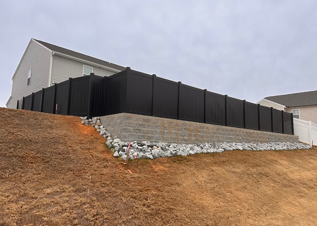 Black vinyl privacy fence installed atop a gray stone retaining wall on a sloped yard with two houses in the background under a cloudy sky.