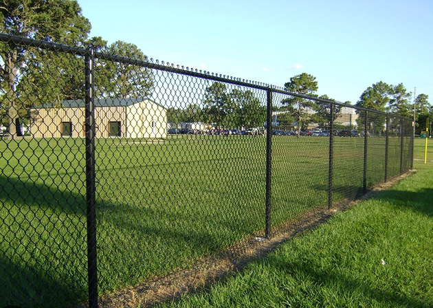 Black chain-link fence enclosing a grassy field with trees and buildings in the background under a clear blue sky.