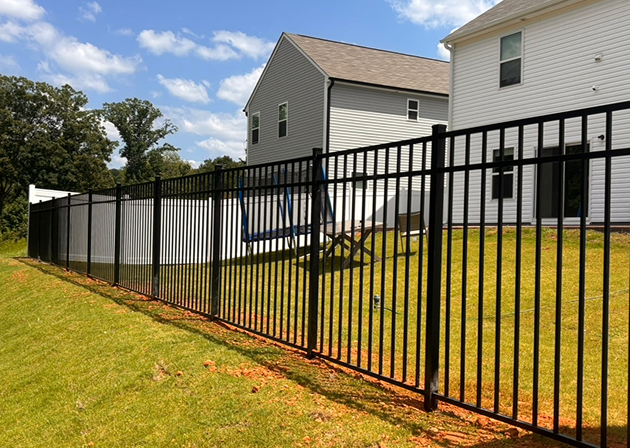 Black metal fence enclosing a grassy yard with two white houses and a blue swing set under a partly cloudy sky.