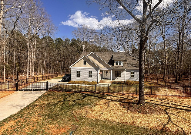 Modern two-story house with light gray siding, a dark roof, and a black metal fence enclosing the front yard with a driveway on the left.