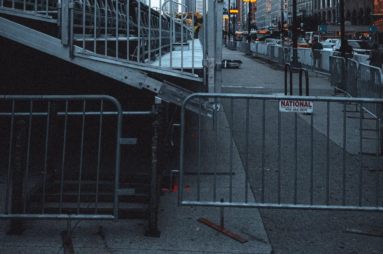 Metal barricades blocking a sidewalk next to a metal staircase in an urban setting with city buildings and traffic lights in the background.