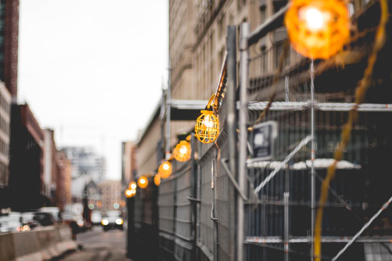 String of yellow construction cage lights along a metal fence on an urban street with blurred buildings and vehicles in the background.