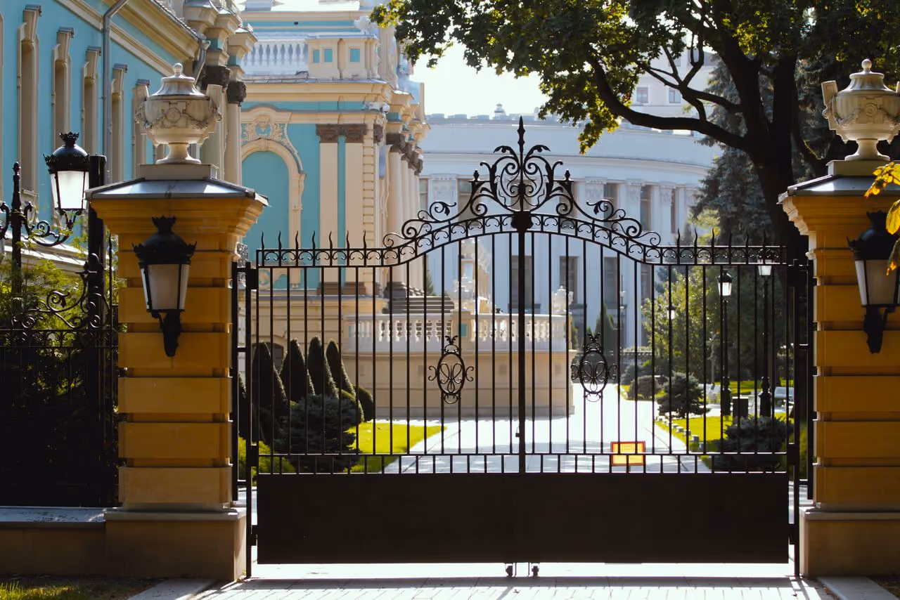 Ornate black wrought iron gate with intricate designs, flanked by tall yellow stone pillars with decorative urns and lamps, opening to a manicured garden and classical architecture buildings in the background under sunlight.