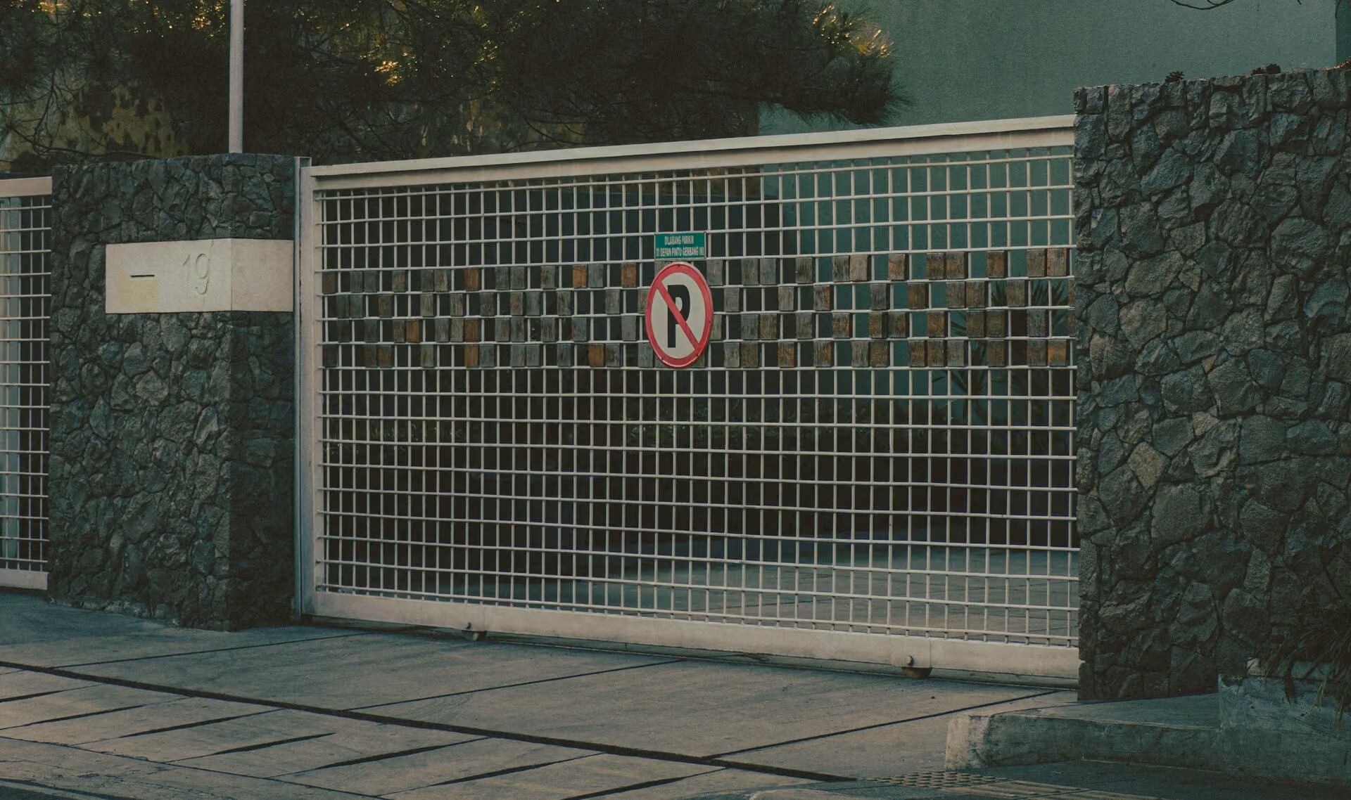 Metal grid gate with a no parking sign and stone pillars on each side, next to a sidewalk and street.