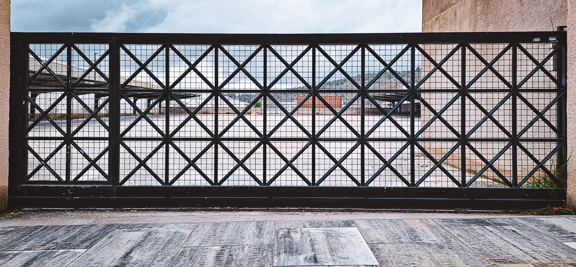 Black metal gate with square and diagonal grid patterns between two beige walls, overlooking an empty paved courtyard with distant hills and cloudy blue sky.