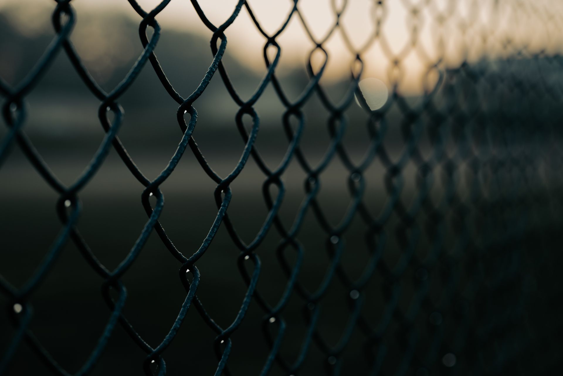 Close-up view of a dark metal chain-link fence with a blurred background at dusk.
