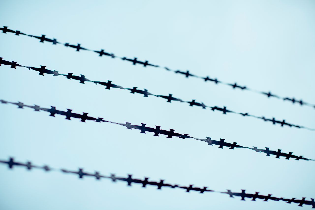 Close-up of several strands of razor wire or barbed tape against a pale blue sky background.