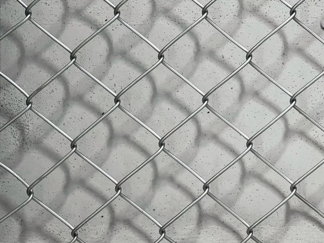 Close-up of a silver chain-link fence casting shadows on a textured gray concrete wall behind it.