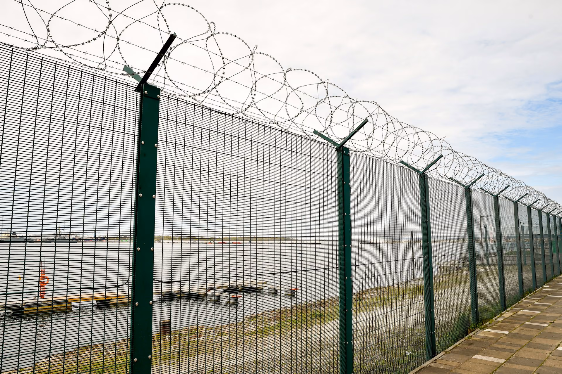 Tall metal security fence topped with spirals of barbed wire along a waterfront with cloudy sky, paved walkway beside the fence.