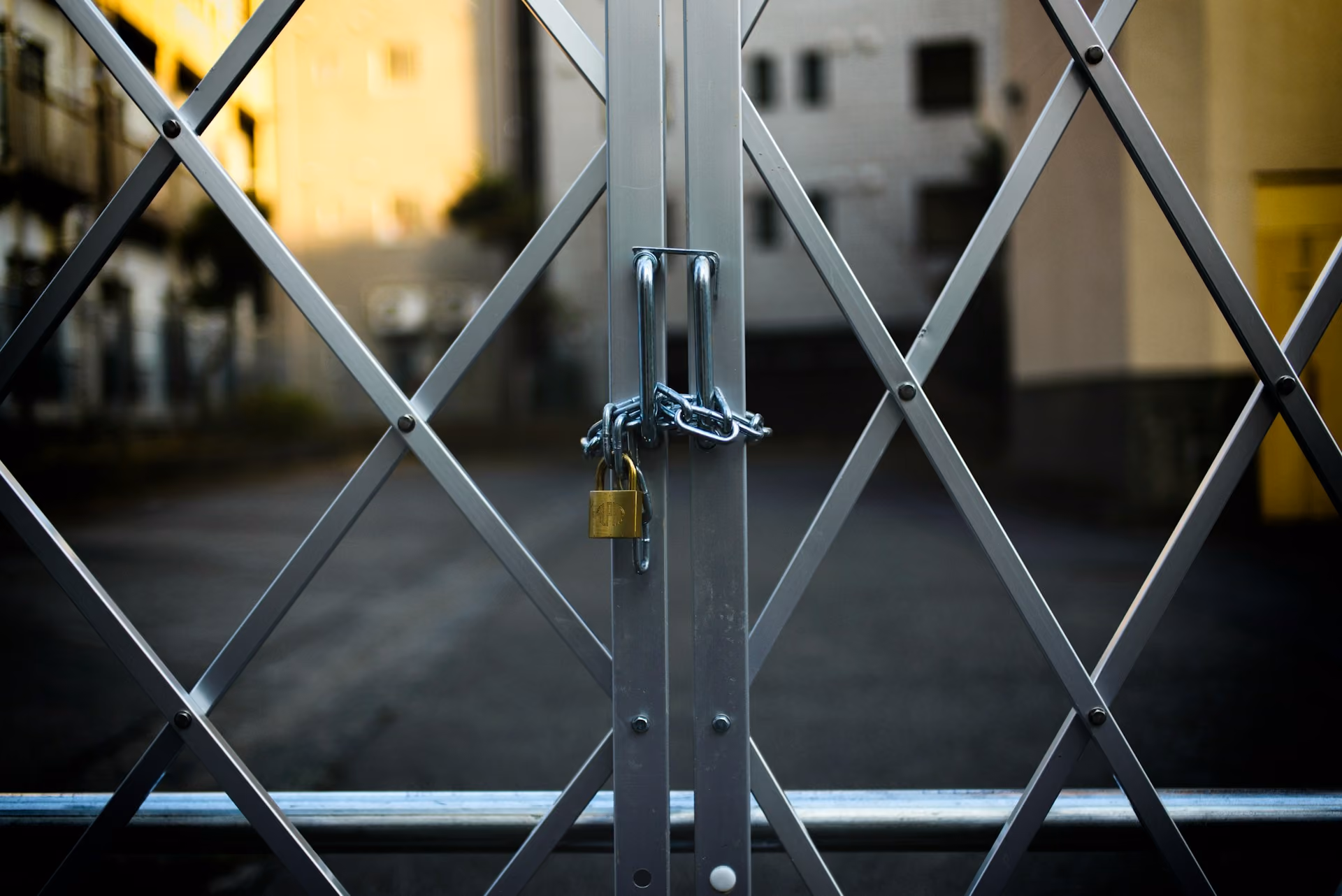 Closed metal gate with a silver chain and a small brass padlock securing the two vertical bars, with blurred background of buildings and pavement.