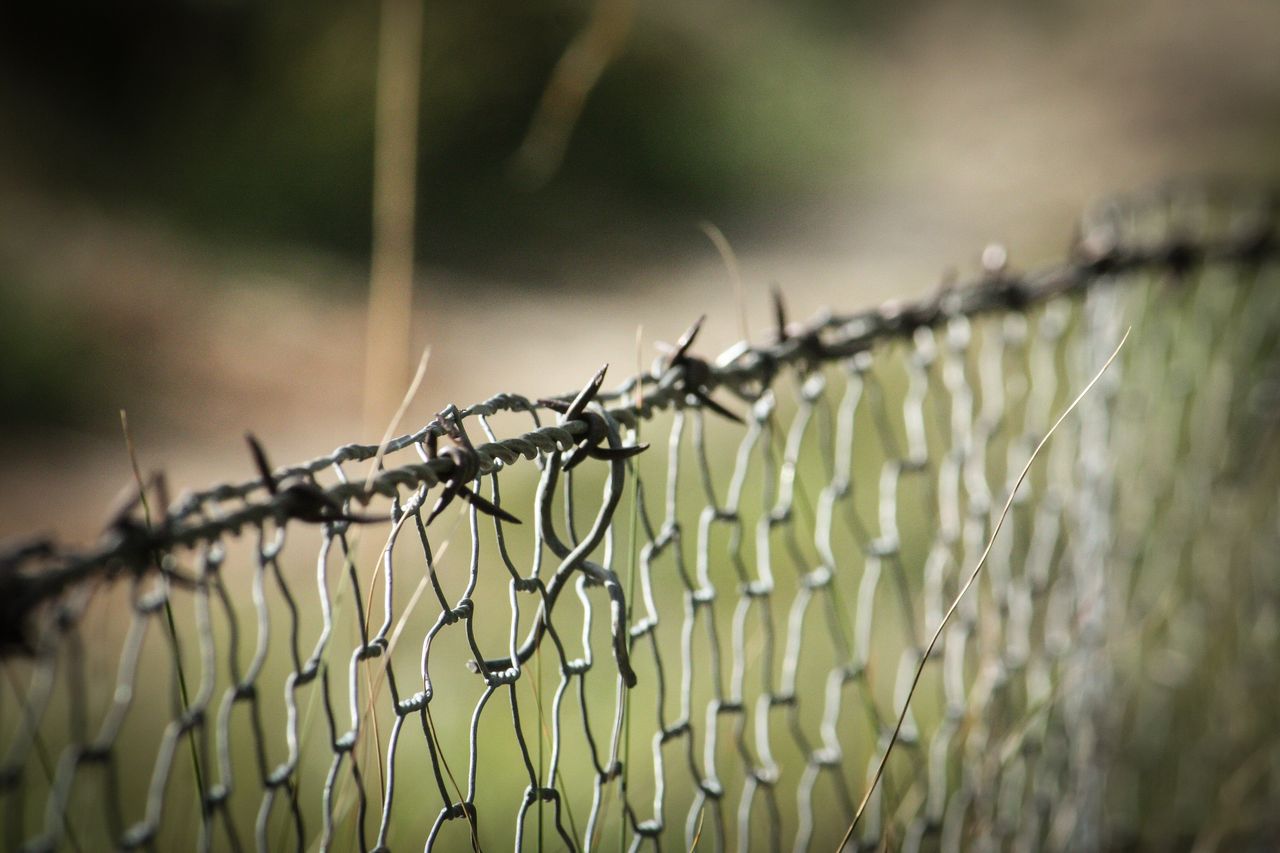 Close-up of a chain-link fence topped with barbed wire, with a blurred natural background.