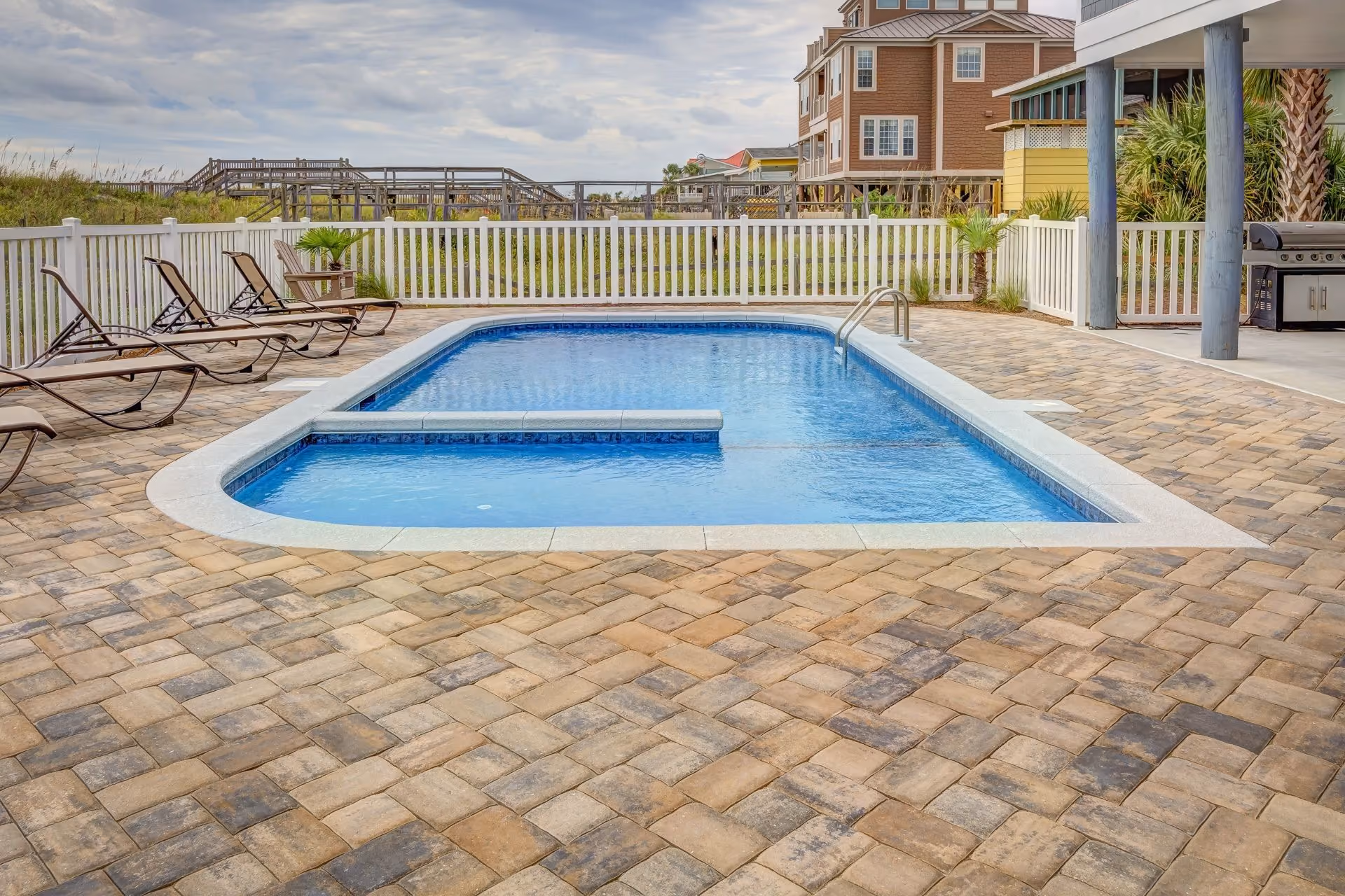 Rectangular outdoor swimming pool with a white border and a dividing ledge in the water, surrounded by tan and brown stone tiles, black metal lounge chairs on the left, white fence enclosing the area, and buildings and greenery in the background under a partly cloudy sky.