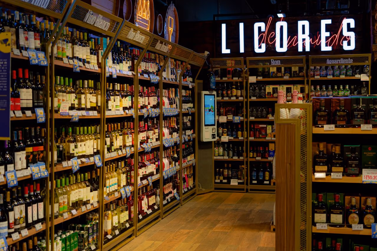 Interior of a liquor store aisle with shelves stocked with various bottles of wine and spirits under illuminated signs for different types like Vino Blanco, Ron, and Aguardiente.