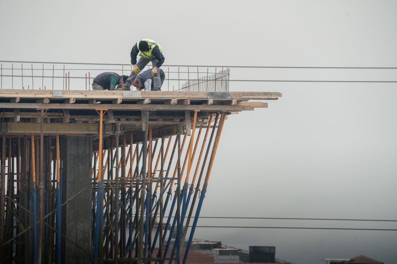 Three construction workers on a partially built concrete structure supported by angled metal scaffolding poles, working under an overcast sky.
