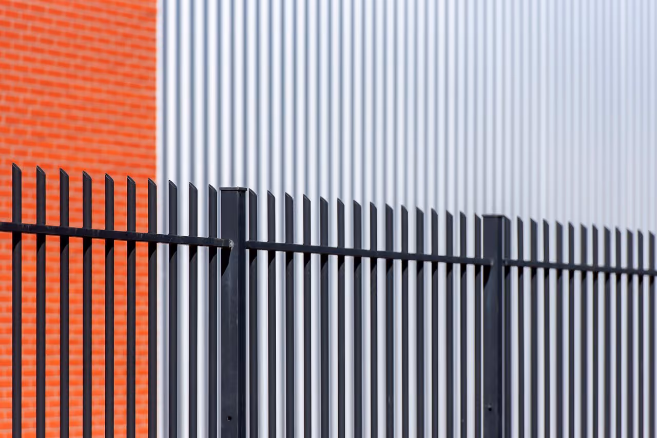 Black metal fence with vertical bars in front of a background split between an orange brick wall and a gray corrugated metal wall.