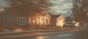 Blurred image of a suburban house with a front porch and garage, with trees and cars nearby.