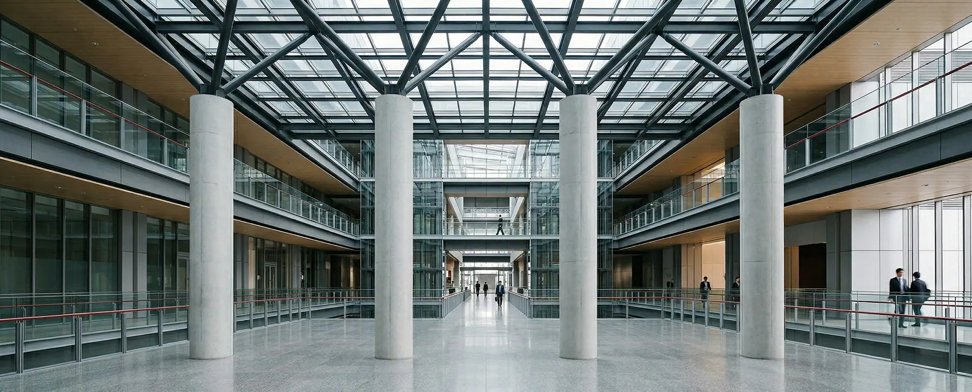 Großer, moderner Innenraum mit vier großen Betonsäulen, Glasgeländern und einem Glasdach mit Stahlträgern.