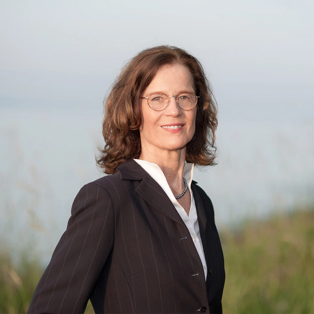 Porträt einer lächelnden Frau mit braunen, welligen Haaren, Brille und schwarzem Nadelstreifenanzug vor verschwommenem Naturhintergrund.