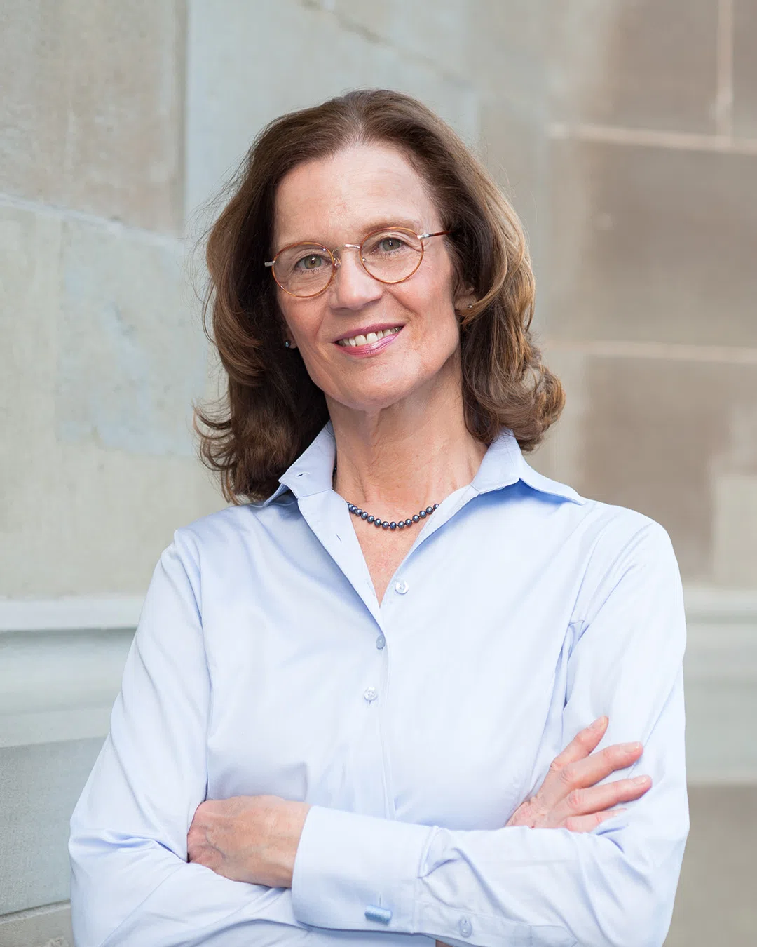 Ältere Frau (Christina Blake) mit Brille und Perlenkette, die in einem hellblauen Hemd lächelnd die Arme verschränkt vor einer Steinwand steht.