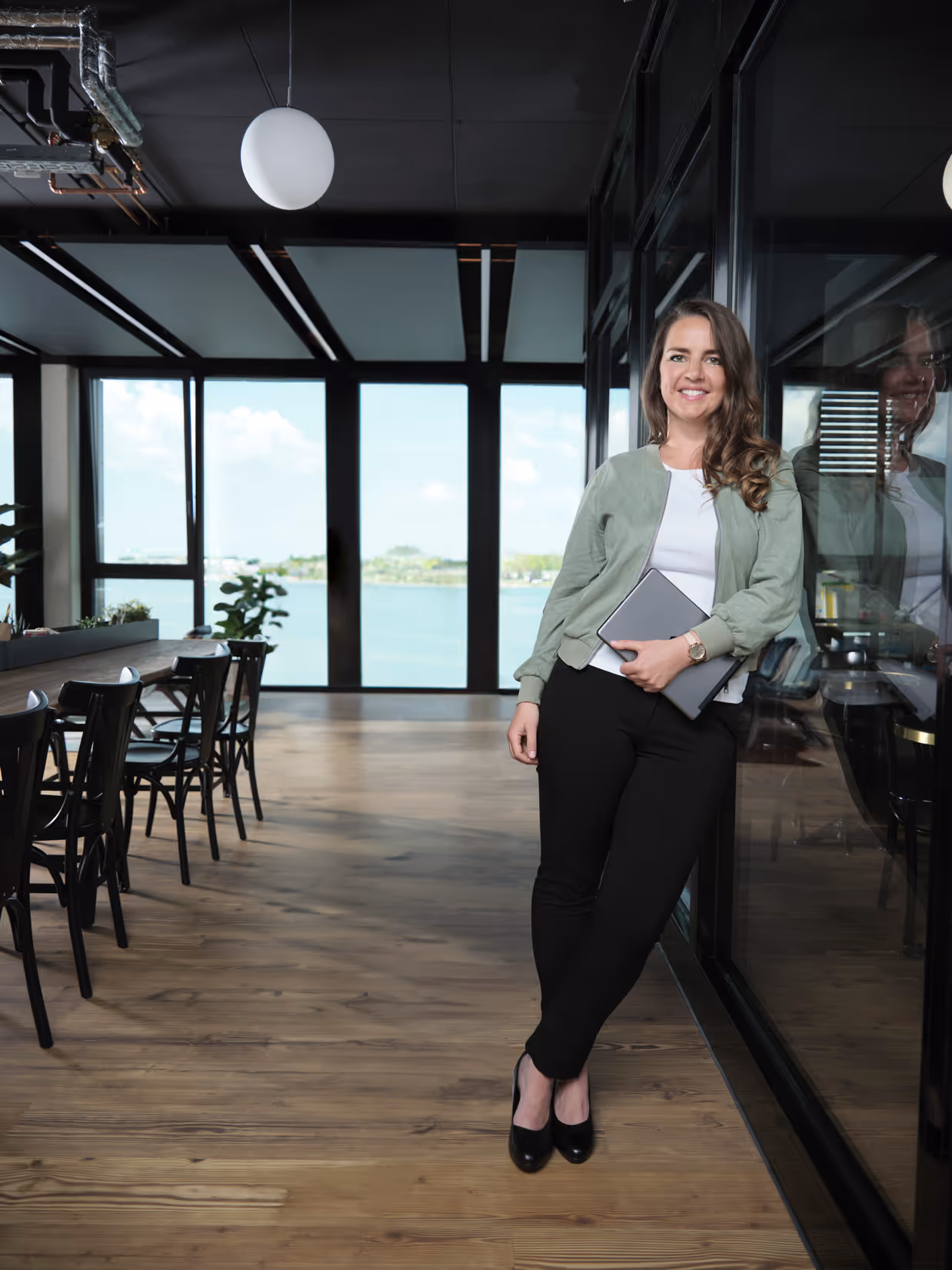 Junge Frau lehnt lächelnd an einer Glaswand in einem modernen Büro mit Blick auf Wasser, hält einen Laptop.