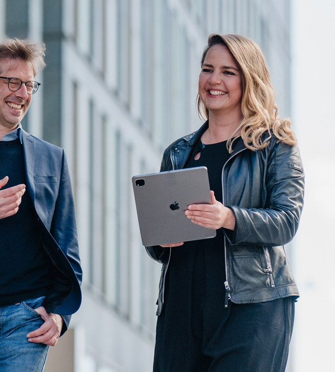 Zwei lächelnde Personen, eine Frau mit Tablet und ein Mann in Brille und Jacke, gehen draußen an modernen Gebäuden entlang.