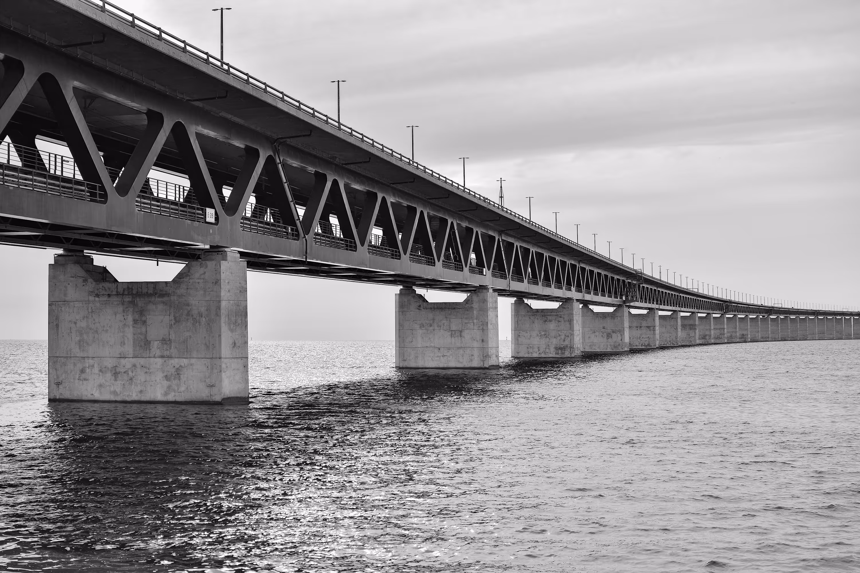 Black and white photo of a long bridge with triangular truss supports extending over calm water under a cloudy sky.