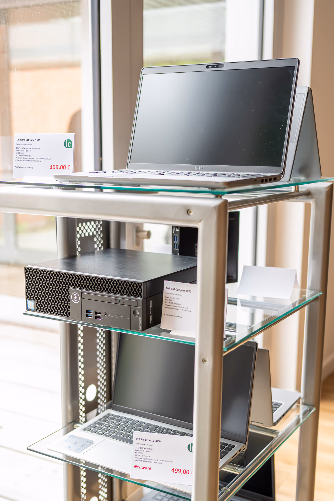 Drei Dell-Computer auf Glasregalen in einem Laden, zwei Laptops und eine Desktop-Einheit mit Preisschildern in Euro.