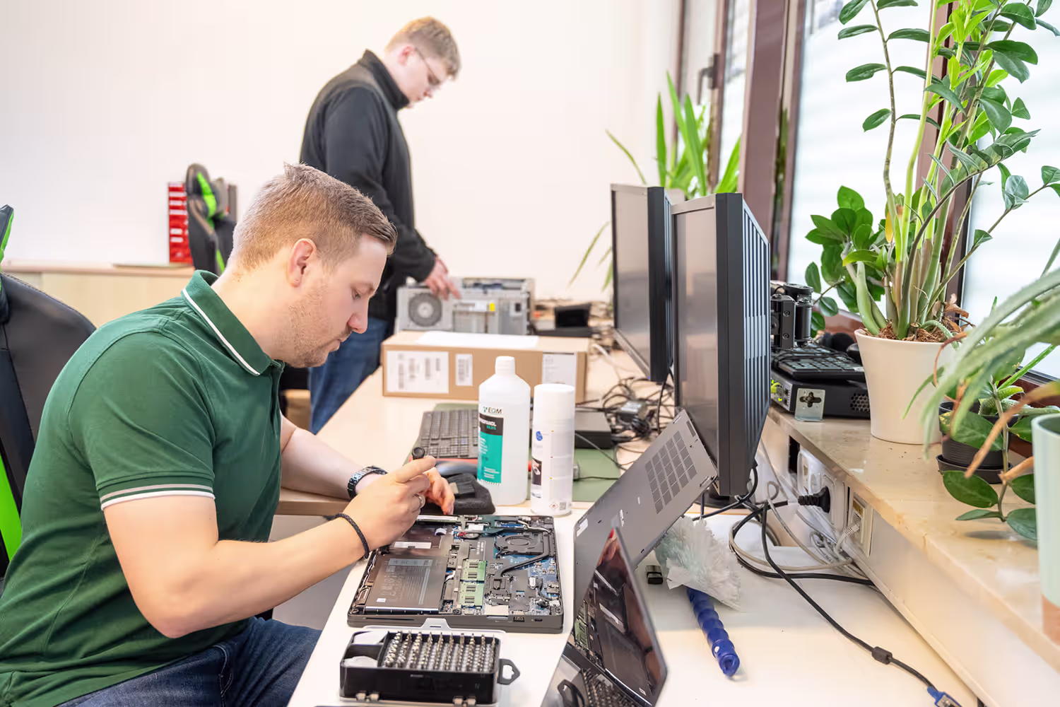 Zwei Männer reparieren Elektronikgeräte an einem Tisch mit geöffnetem Laptop, Werkzeug und Pflanzen am Fenster.