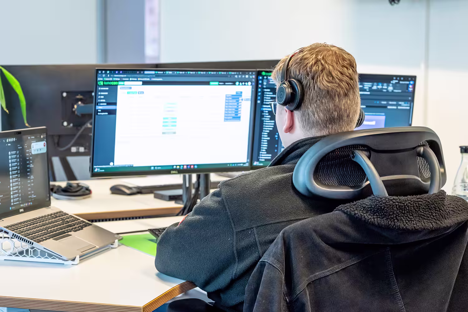 Person sitzt an einem Schreibtisch mit zwei Monitoren und einem Laptop und trägt Kopfhörer.