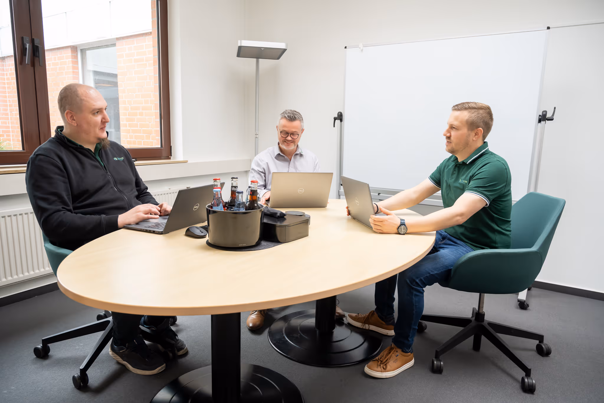 Drei Männer sitzen an einem ovalen Konferenztisch mit Laptops und Getränken in einem hellen Raum mit Whiteboard.