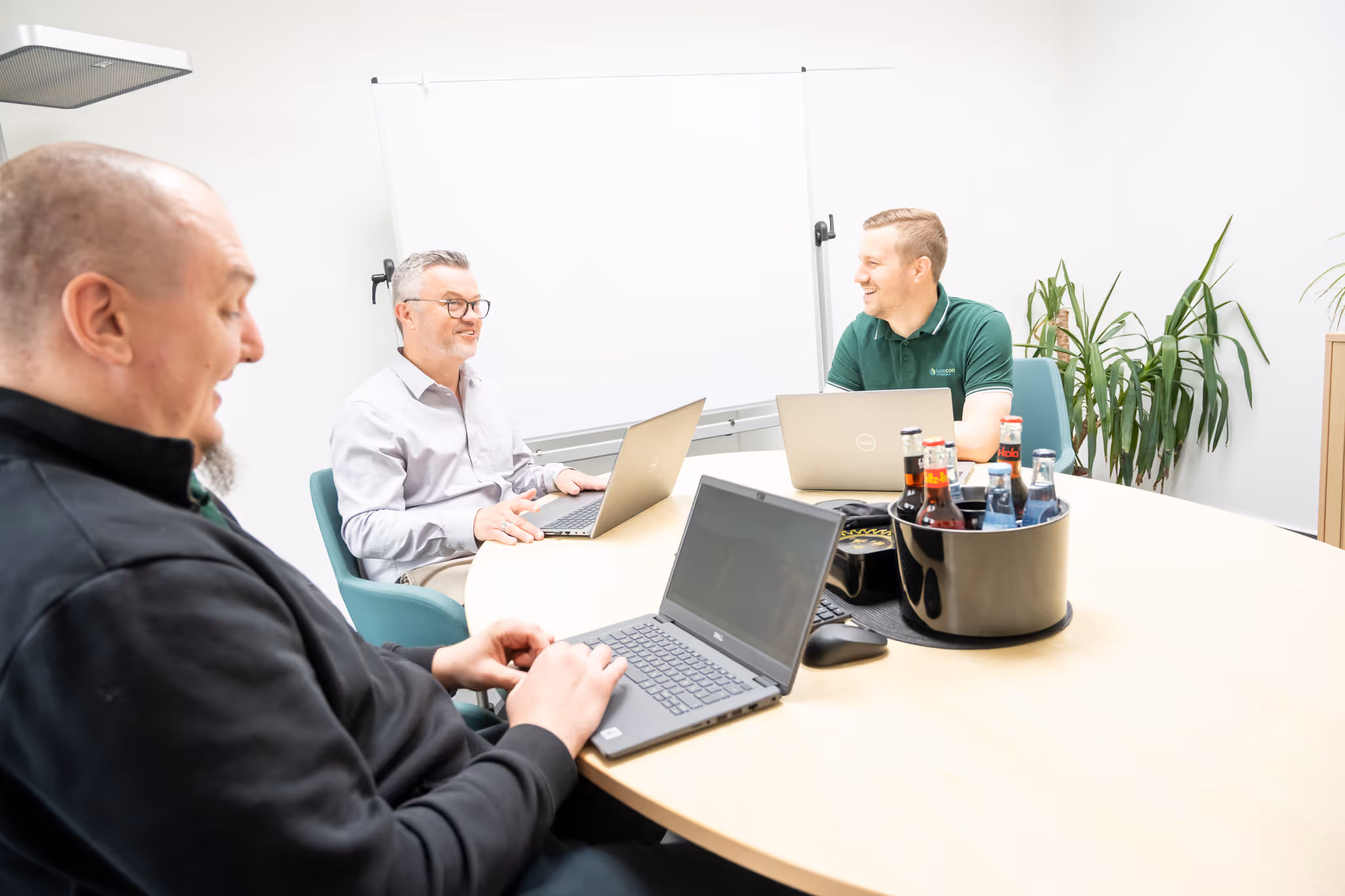 Drei Männer sitzen an einem runden Tisch mit Laptops und unterhalten sich in einem hellen Besprechungsraum.