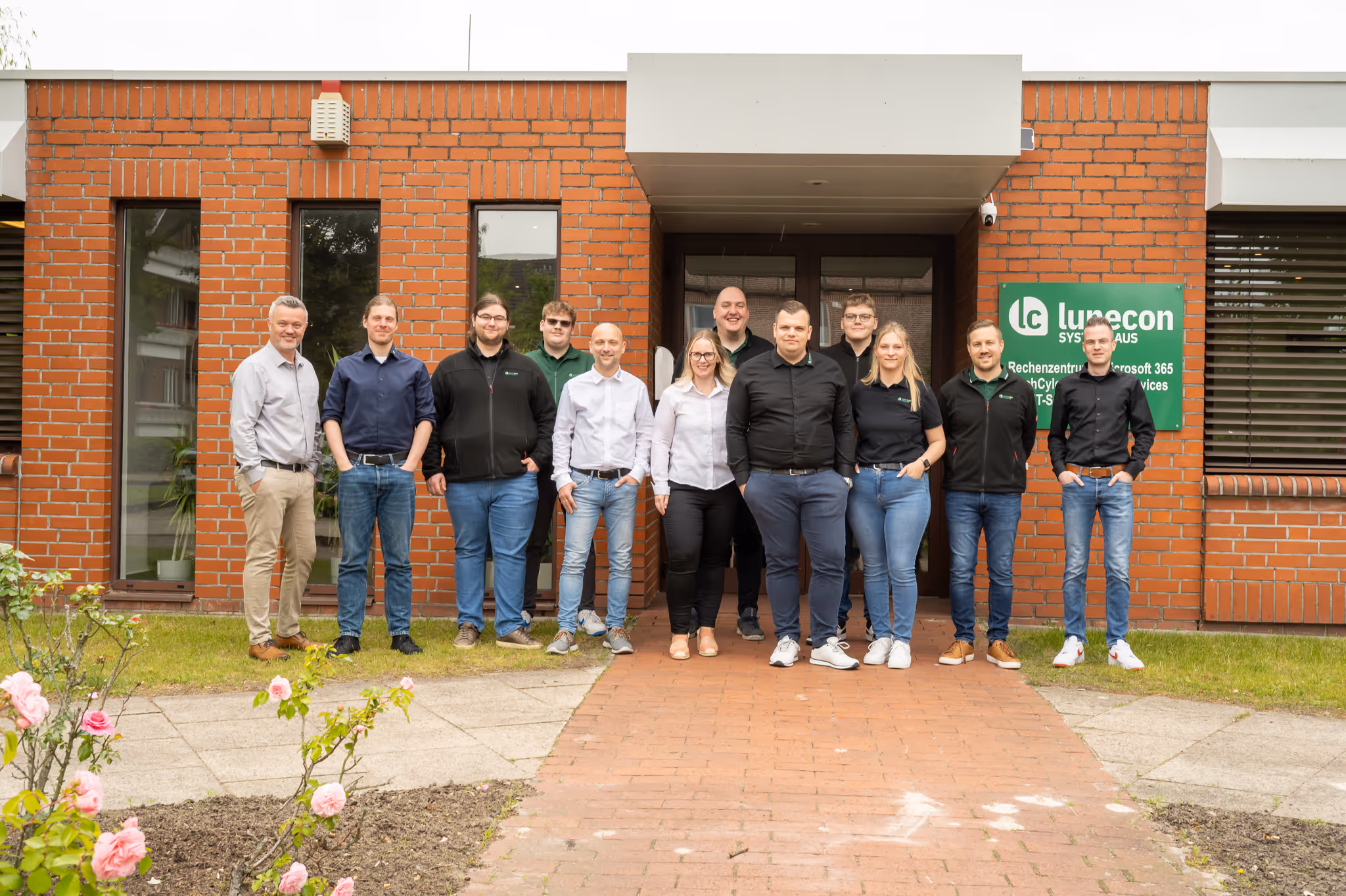 Gruppenfoto von elf Personen, die vor einem roten Backsteingebäude mit einem Eingang stehen, auf dem ein grünes Lunecon-Schild angebracht ist.