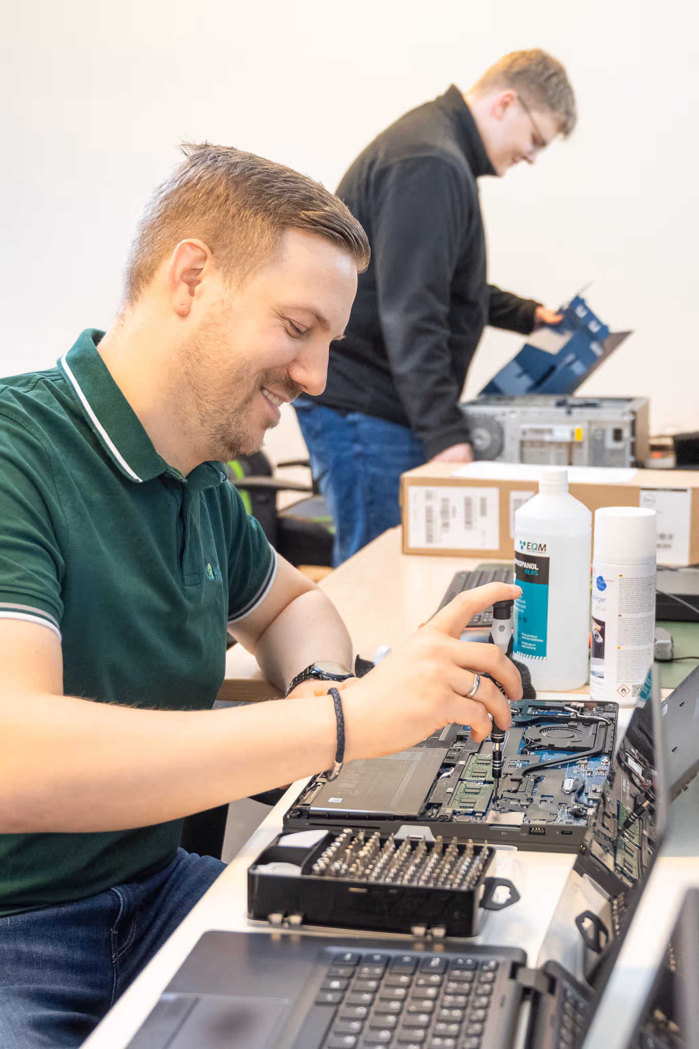 Techniker repariert lächelnd einen geöffneten Laptop, während eine andere Person im Hintergrund arbeitet.