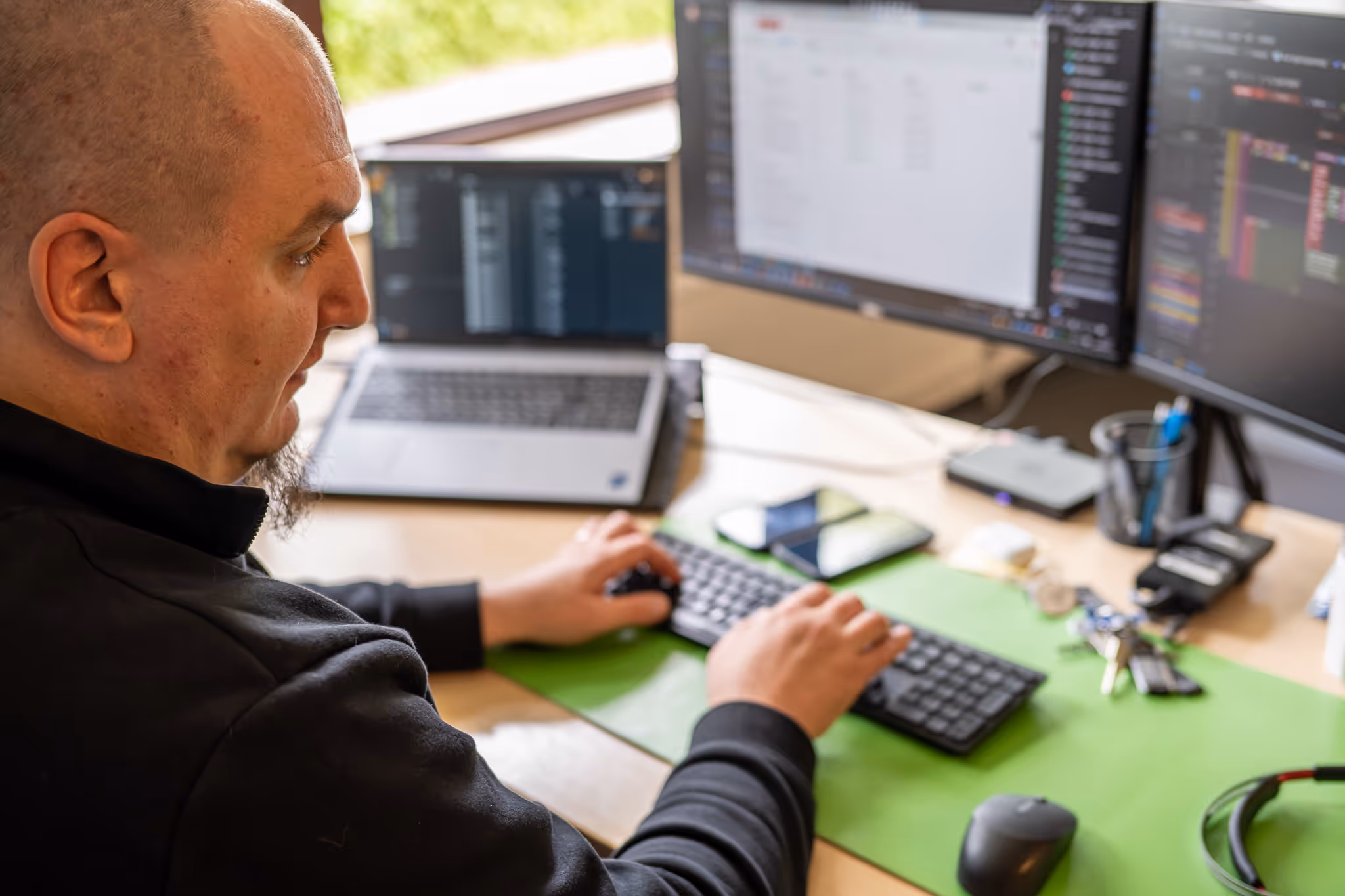 Ein Mann tippt auf einer Tastatur vor zwei Monitoren und einem Laptop auf einem Schreibtisch mit verschiedenen Accessoires.