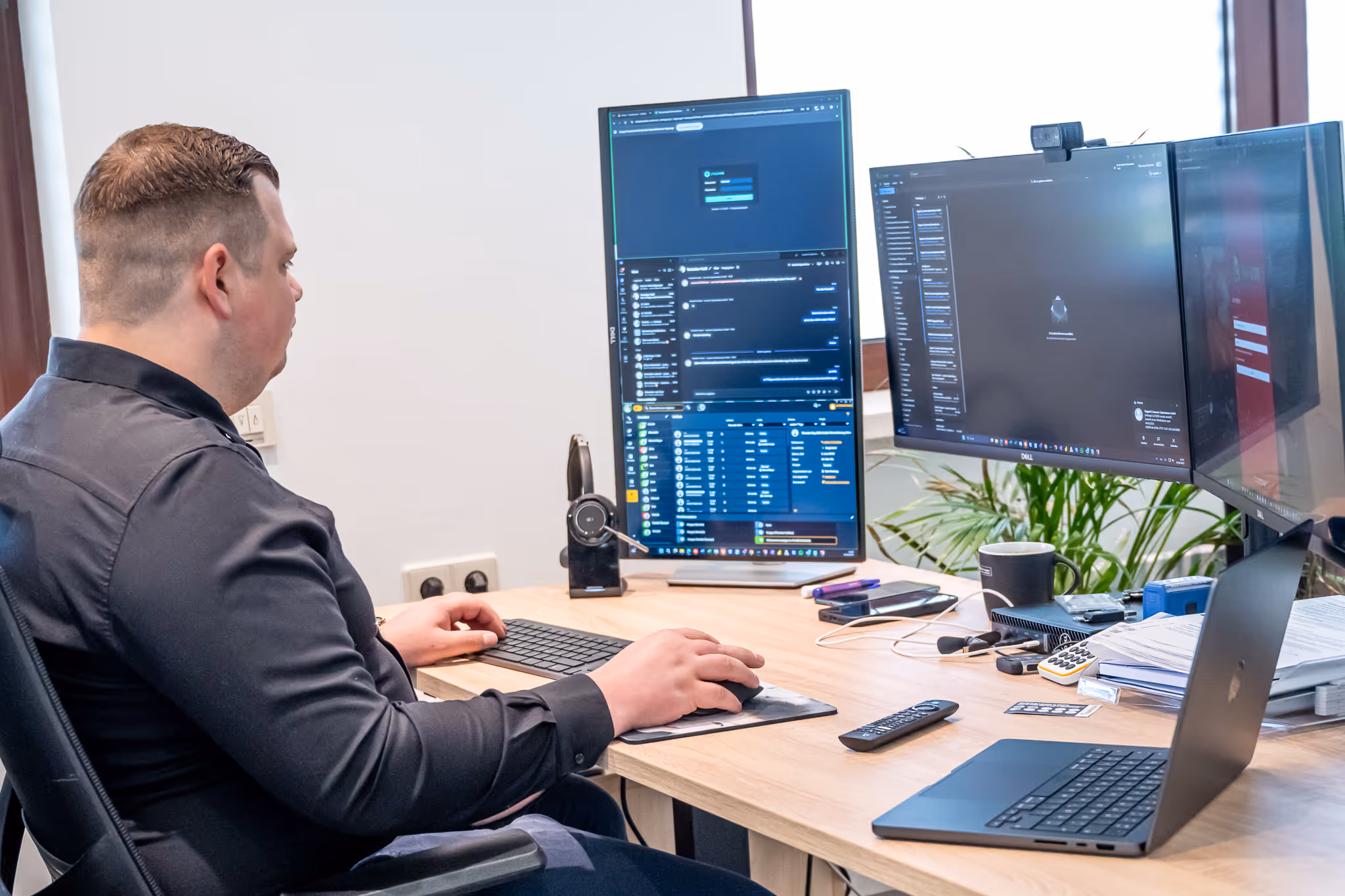 Ein Mann sitzt am Schreibtisch und arbeitet an einem Computer mit drei Monitoren und einem Laptop.