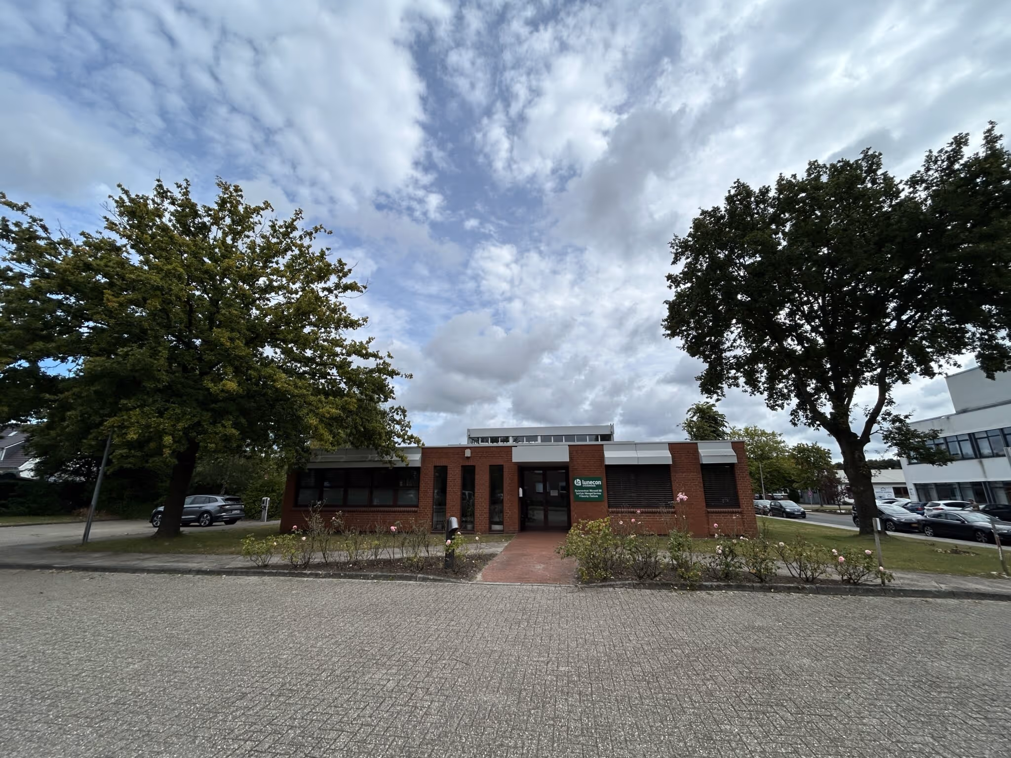 Ein einstöckiges Backsteingebäude mit einem begrünten Vorgarten und zwei großen Bäumen auf einer gepflasterten Straße unter bewölktem Himmel.