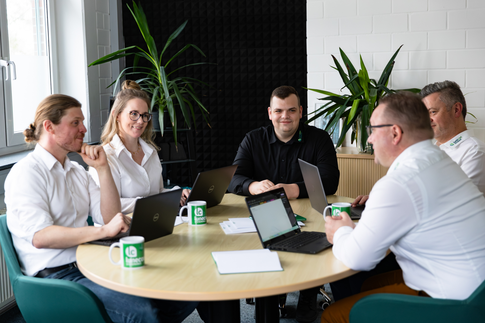Fünf Personen in weißem und schwarzem Hemd sitzen an einem runden Tisch mit Laptops und grünen Kaffeetassen in einem modernen Büro.