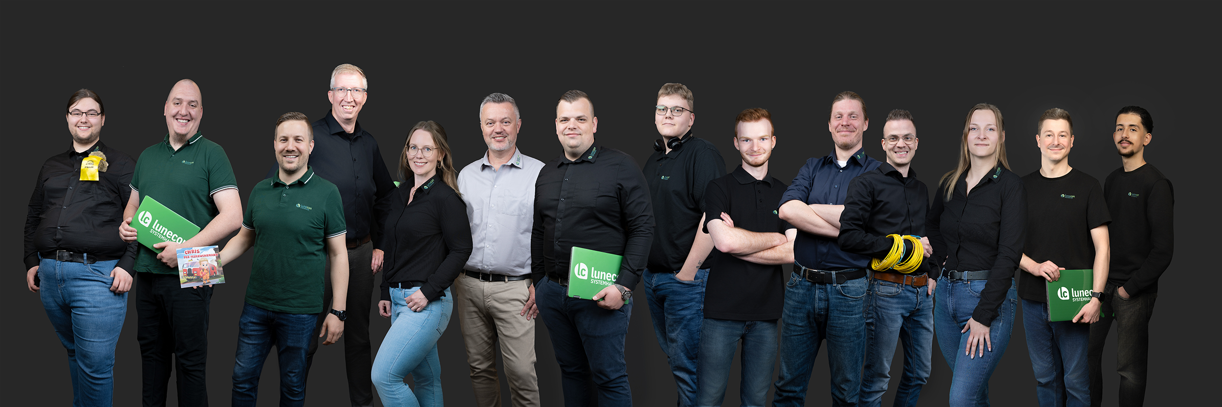 Gruppenfoto von zwölf Mitarbeitern des Unternehmen Lunecon Systemhaus vor dunklem Hintergrund, einige halten grüne Laptops mit dem Firmenlogo.