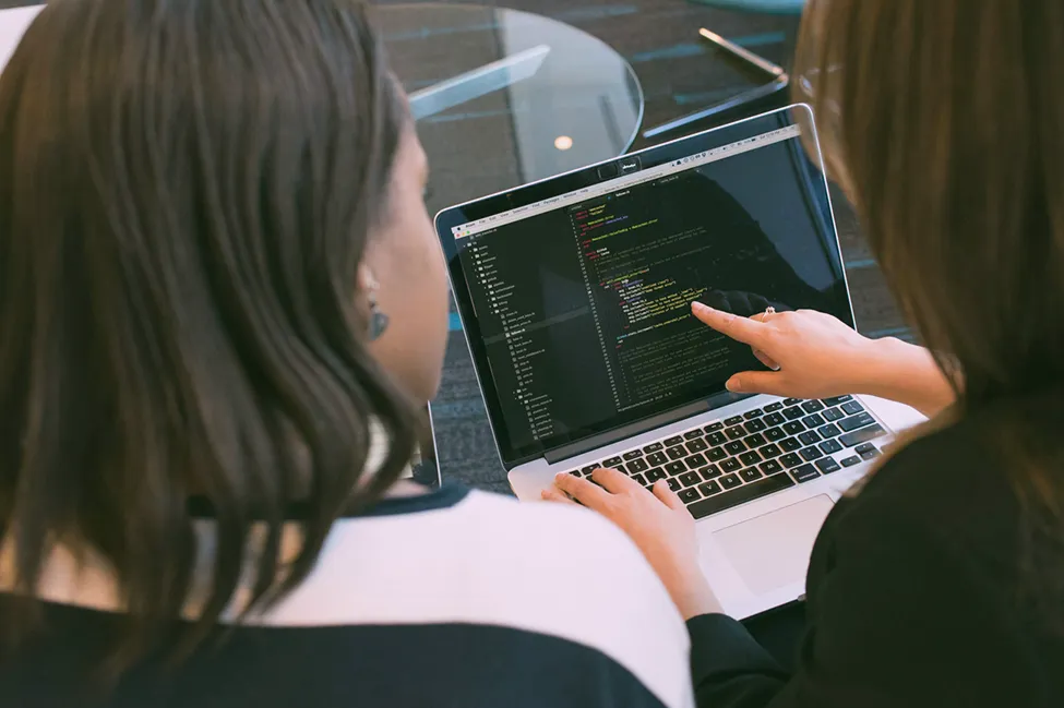 Een foto van 2 vrouwen die achter een laptop met code zitten. Een vrouw wijst naar een lijn code.
