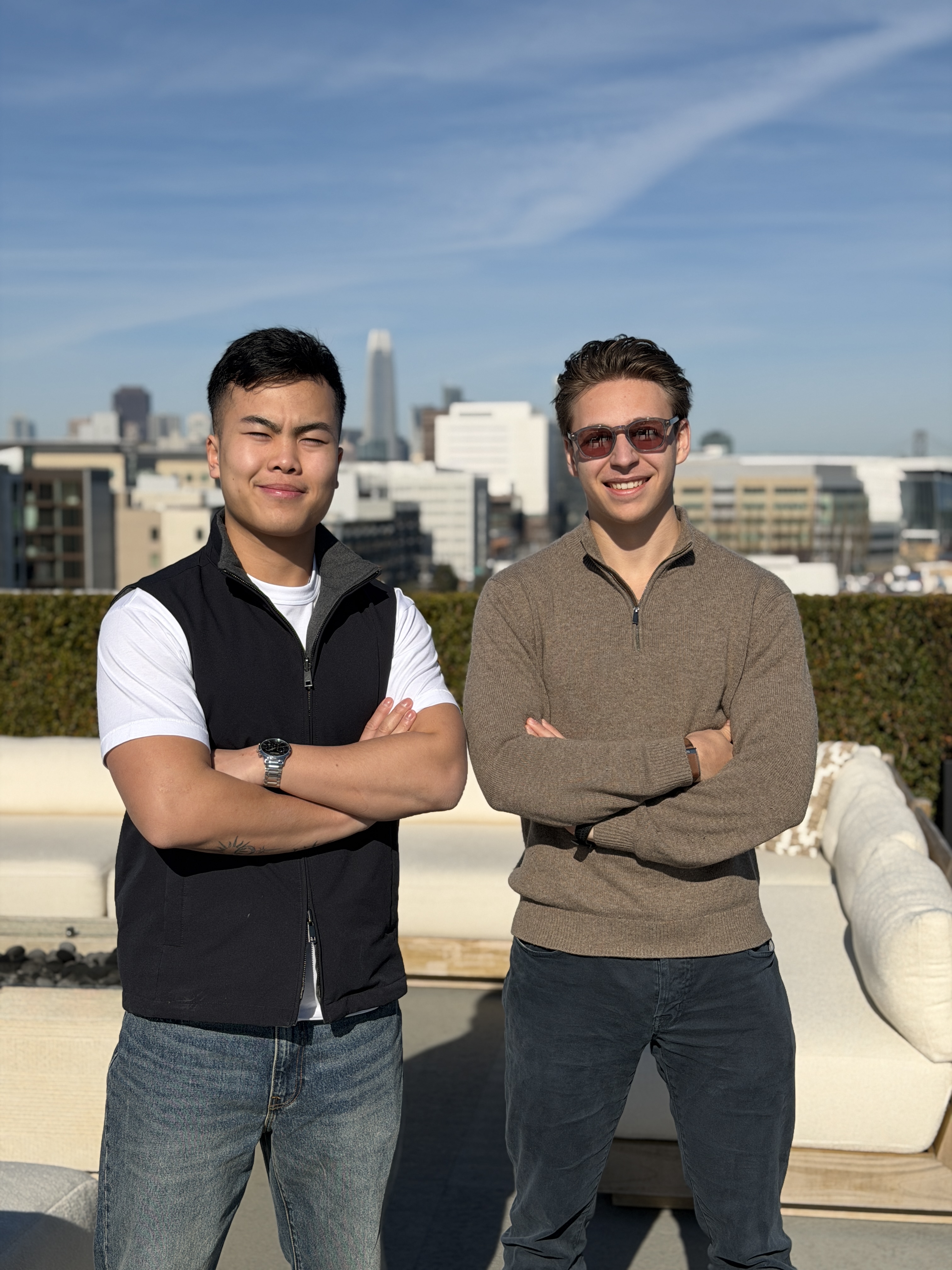 Two young men from Daymaker standing outdoors with arms crossed, smiling in front of a cityscape under a blue sky.