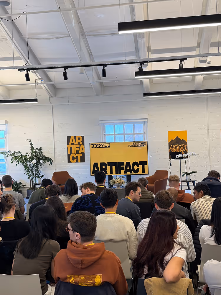 Audience seated in a room facing a stage with yellow banners displaying the word 'ARTIFACT' and event details.