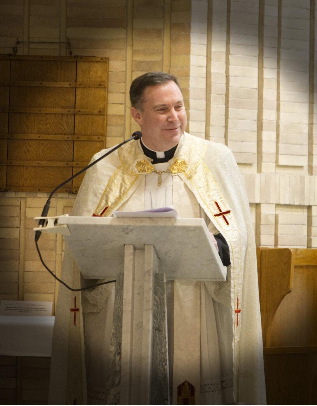 A priest in white and gold vestments standing behind a marble lectern with a microphone, smiling during a church service.