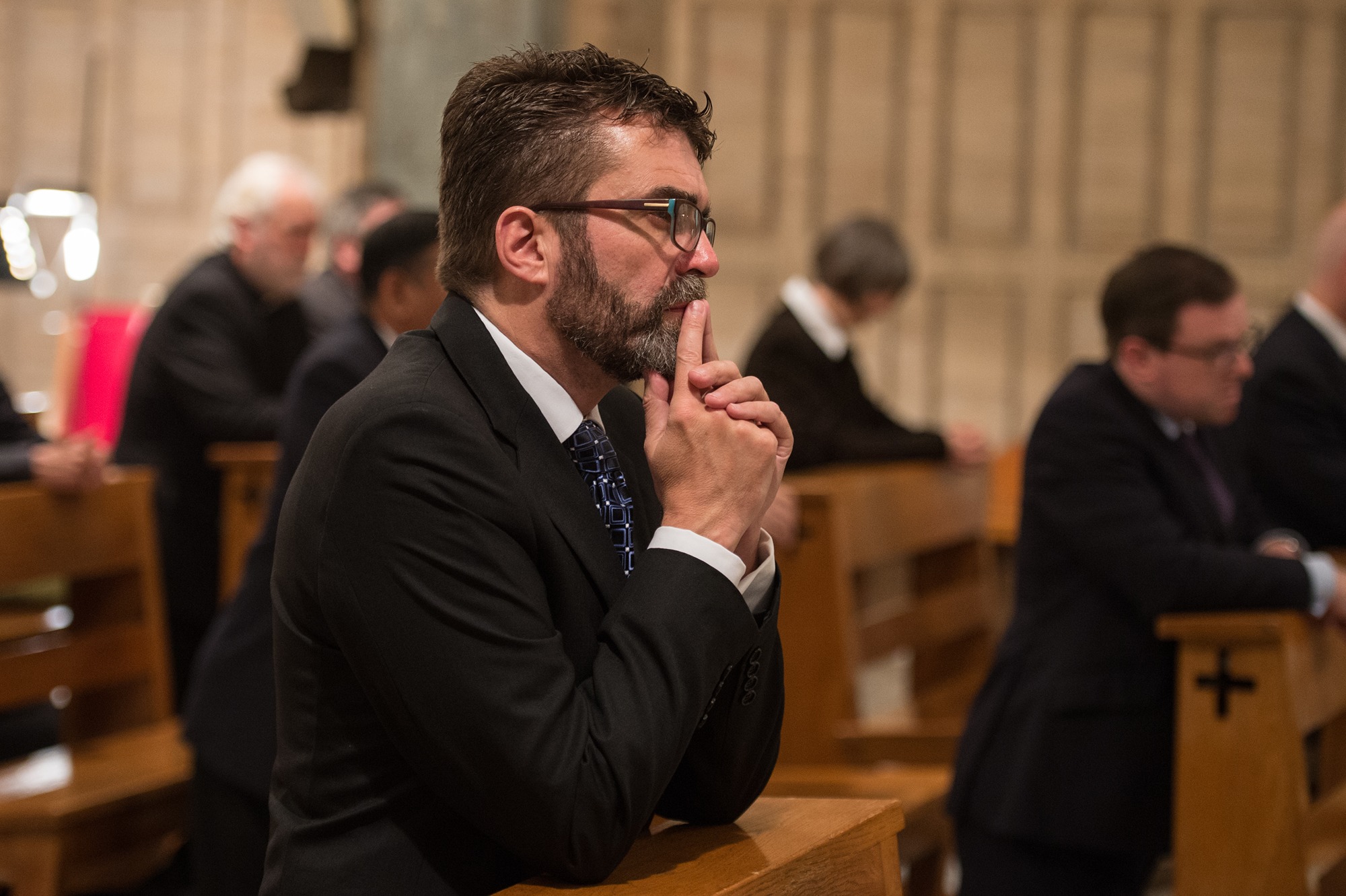 Man in a suit and glasses praying thoughtfully in a church pew with other worshippers in the background.