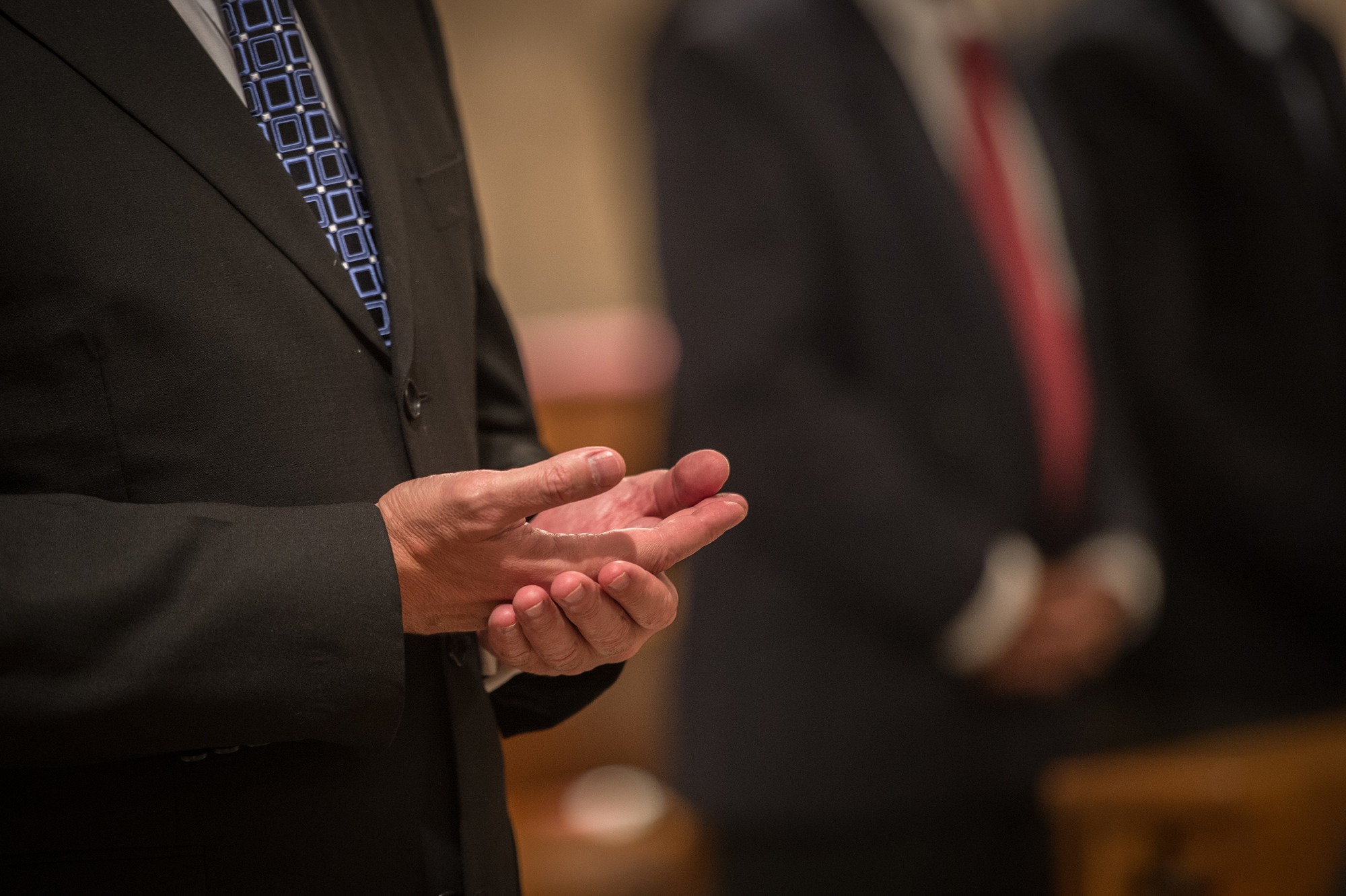 Close-up of a person in a suit with a geometric-patterned tie holding their hands open in front of them, with blurred people in suits in the background.